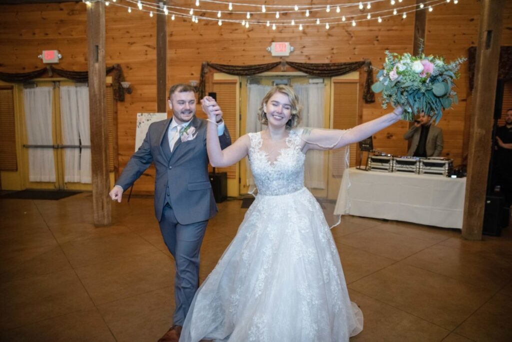 Christine-Jon-Wedding-Blog-93 - TOMMY GATZ ENTERTAINMENT A bride and groom walk hand in hand in a wooden hall. The bride is smiling and holding a bouquet, and the groom is wearing a suit. String lights are hanging above.