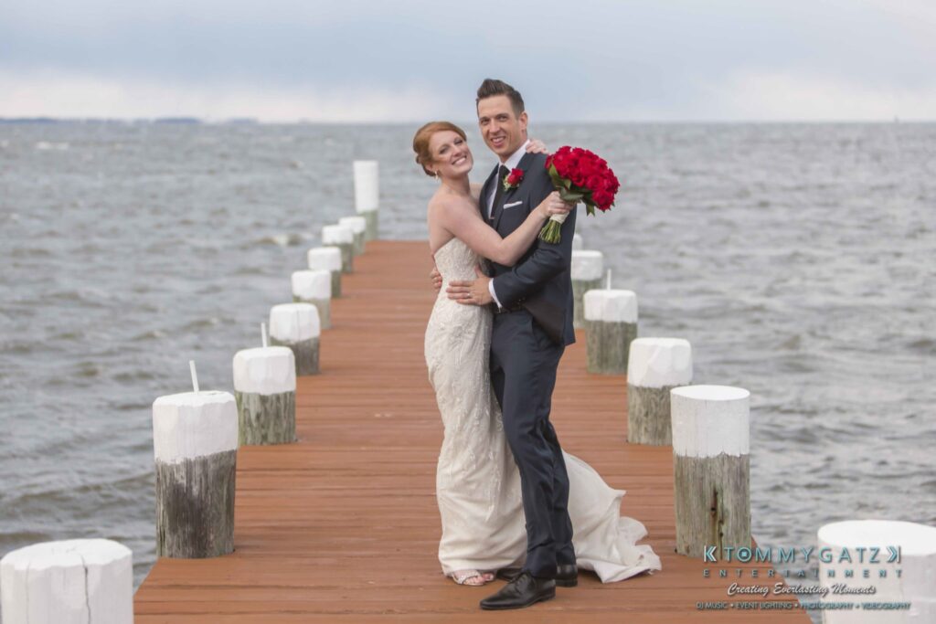 Bride and groom pose on a wooden pier over water, holding a bouquet of red roses.
