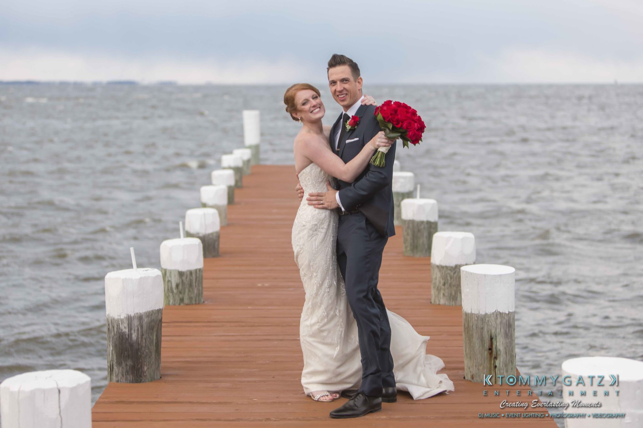 Bride and groom pose on a wooden pier over water, holding a bouquet of red roses.