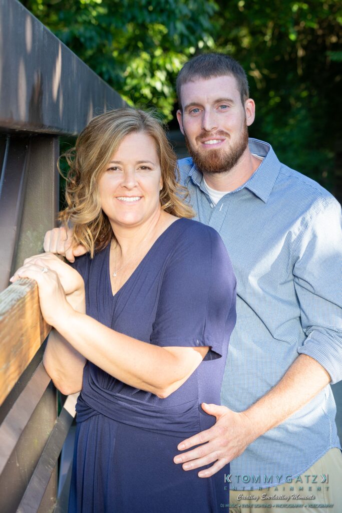 Heather-Scott-Engagement-Shoot-Low-Res-WM-10 - TOMMY GATZ ENTERTAINMENT A woman and a man stand closely together on a bridge, with greenery in the background. The woman leans on the railing while the man stands behind her, resting his hand on her arm.