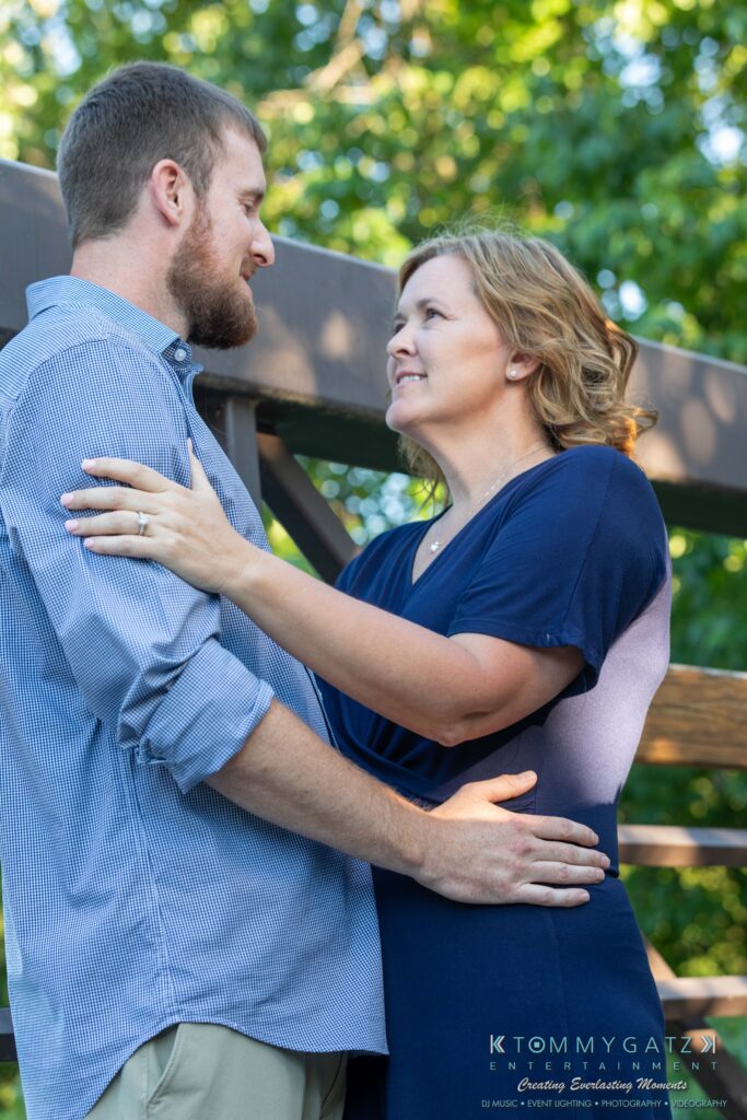 Heather-Scott-Engagement-Shoot-Low-Res-WM-15 - TOMMY GATZ ENTERTAINMENT A man and a woman stand outdoors on a wooden bridge, facing each other and smiling. Trees and sunlight form the background.