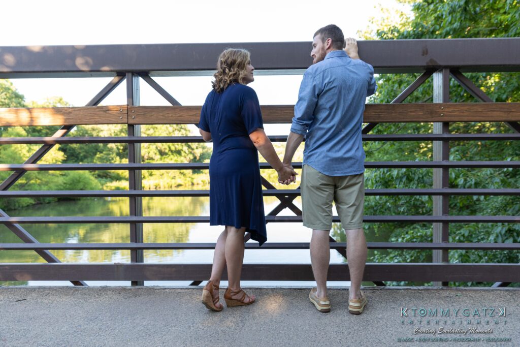 Heather-Scott-Engagement-Shoot-Low-Res-WM-17 - TOMMY GATZ ENTERTAINMENT A couple stands hand in hand on a bridge, looking at each other, with trees and water in the background.