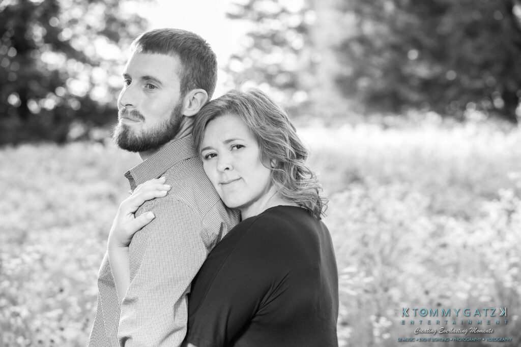 Heather-Scott-Engagement-Shoot-Low-Res-WM-19 - TOMMY GATZ ENTERTAINMENT A woman stands with her arms around a bearded man in an outdoor field setting. Both look in the same direction with neutral expressions. The scene is in black and white.