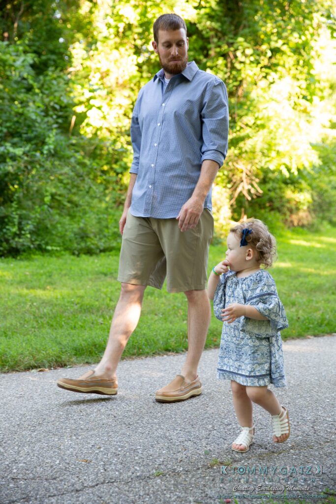 Heather-Scott-Engagement-Shoot-Low-Res-WM-2 - TOMMY GATZ ENTERTAINMENT A man and a toddler walk on a path in a park. The man wears a blue shirt and shorts, while the child wears a floral dress with a bow in her hair. They are surrounded by greenery.