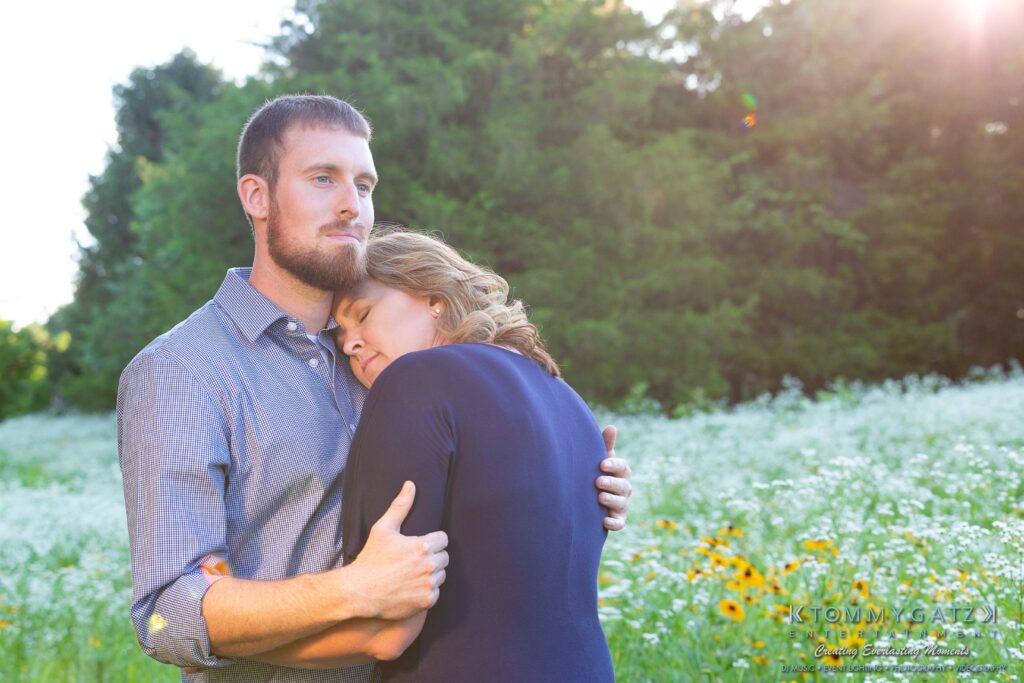 Heather-Scott-Engagement-Shoot-Low-Res-WM-22 - TOMMY GATZ ENTERTAINMENT A couple stands in a field of flowers, with the woman resting her head on the man's chest. Sunlight filters through the trees in the background.