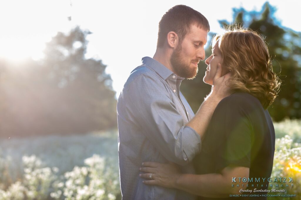Heather-Scott-Engagement-Shoot-Low-Res-WM-28 - TOMMY GATZ ENTERTAINMENT A couple stands closely in a sunlit field, embracing gently, with one person touching the other's face.