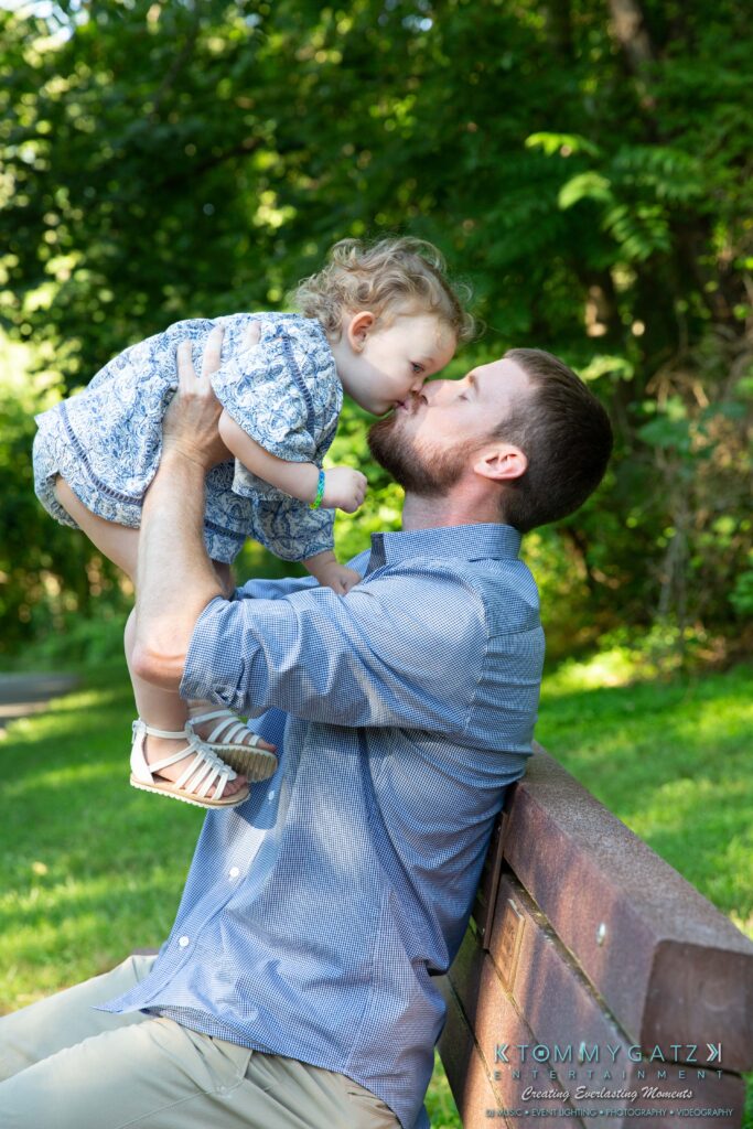 Heather-Scott-Engagement-Shoot-Low-Res-WM-3 - TOMMY GATZ ENTERTAINMENT Man lifting a child over his head, sharing a nose-to-nose moment on a park bench with trees in the background.
