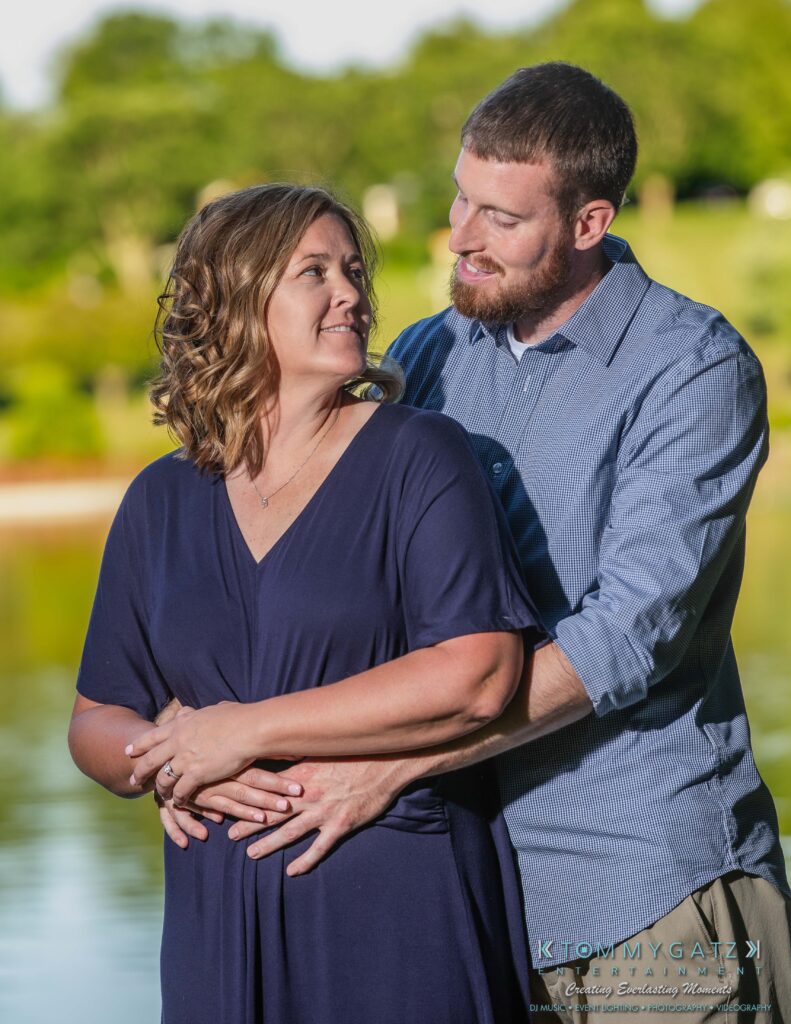 Heather-Scott-Engagement-Shoot-Low-Res-WM-30 - TOMMY GATZ ENTERTAINMENT A couple stands by a lake, with the man embracing the woman from behind. Both are smiling at each other, surrounded by greenery.