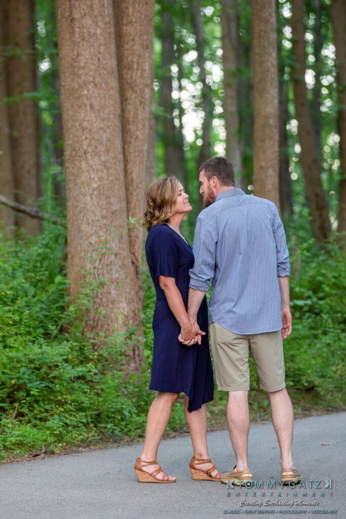 Heather-Scott-Engagement-Shoot-Low-Res-WM-33 - TOMMY GATZ ENTERTAINMENT A couple holding hands, with their foreheads touching, stands on a forest path surrounded by tall trees and greenery.