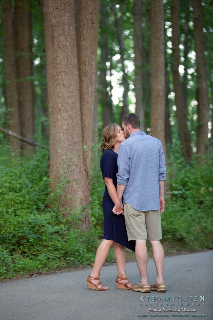 Heather-Scott-Engagement-Shoot-Low-Res-WM-34 - TOMMY GATZ ENTERTAINMENT A couple holding hands, walking on a forest path, both in casual summer clothes. The forest is lush and green.
