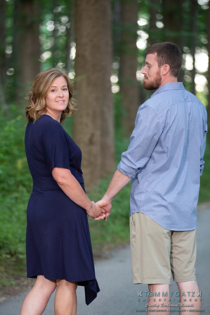 Heather-Scott-Engagement-Shoot-Low-Res-WM-35 - TOMMY GATZ ENTERTAINMENT A couple holds hands while walking on a forested path. The woman looks back at the camera.