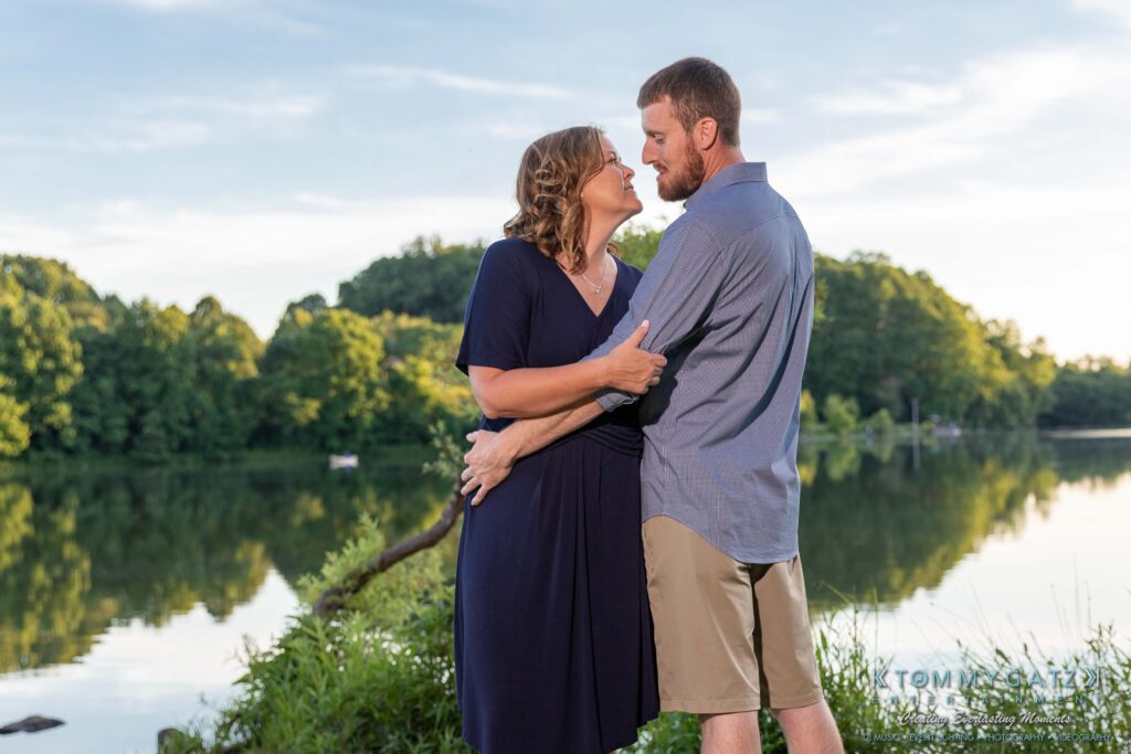 Heather-Scott-Engagement-Shoot-Low-Res-WM-36 - TOMMY GATZ ENTERTAINMENT A couple stands near a lake, embracing and about to kiss, with trees and a clear sky in the background.