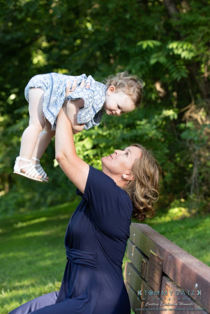 Heather-Scott-Engagement-Shoot-Low-Res-WM-4 - TOMMY GATZ ENTERTAINMENT A woman sitting on a bench lifts a child in a dress above her head. They are outdoors surrounded by greenery.