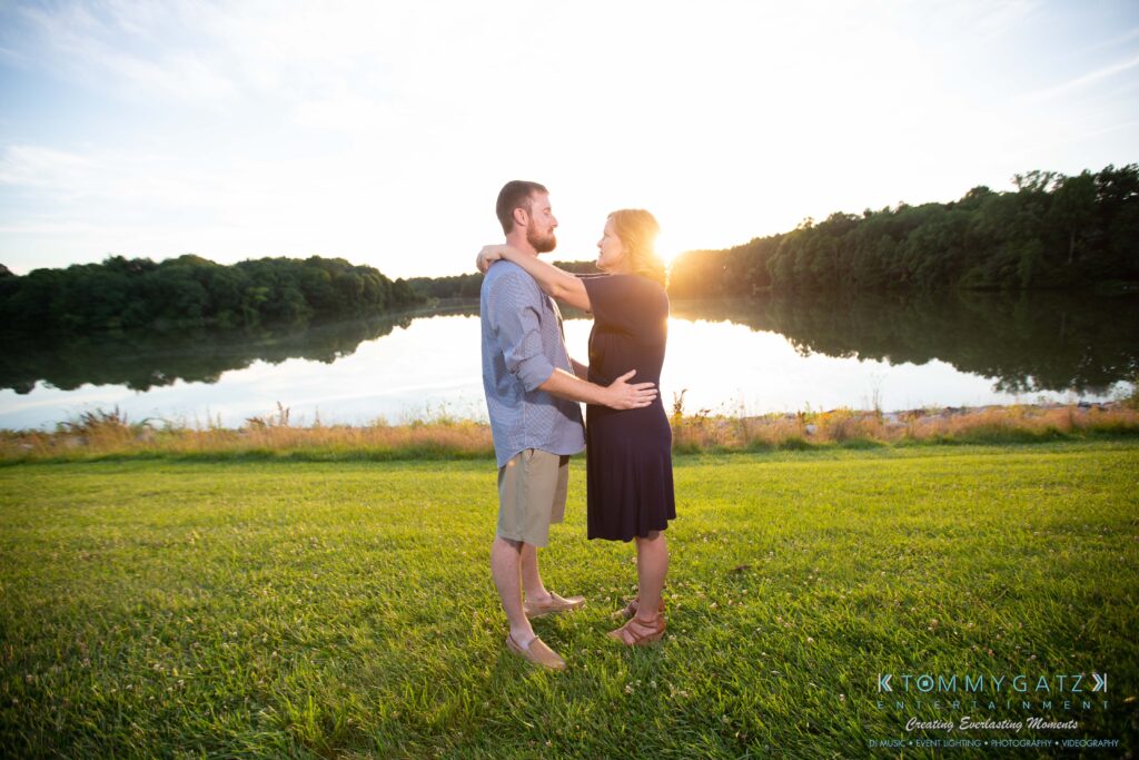 Heather-Scott-Engagement-Shoot-Low-Res-WM-41 - TOMMY GATZ ENTERTAINMENT A couple stands facing each other on grass by a lake at sunset, surrounded by trees.