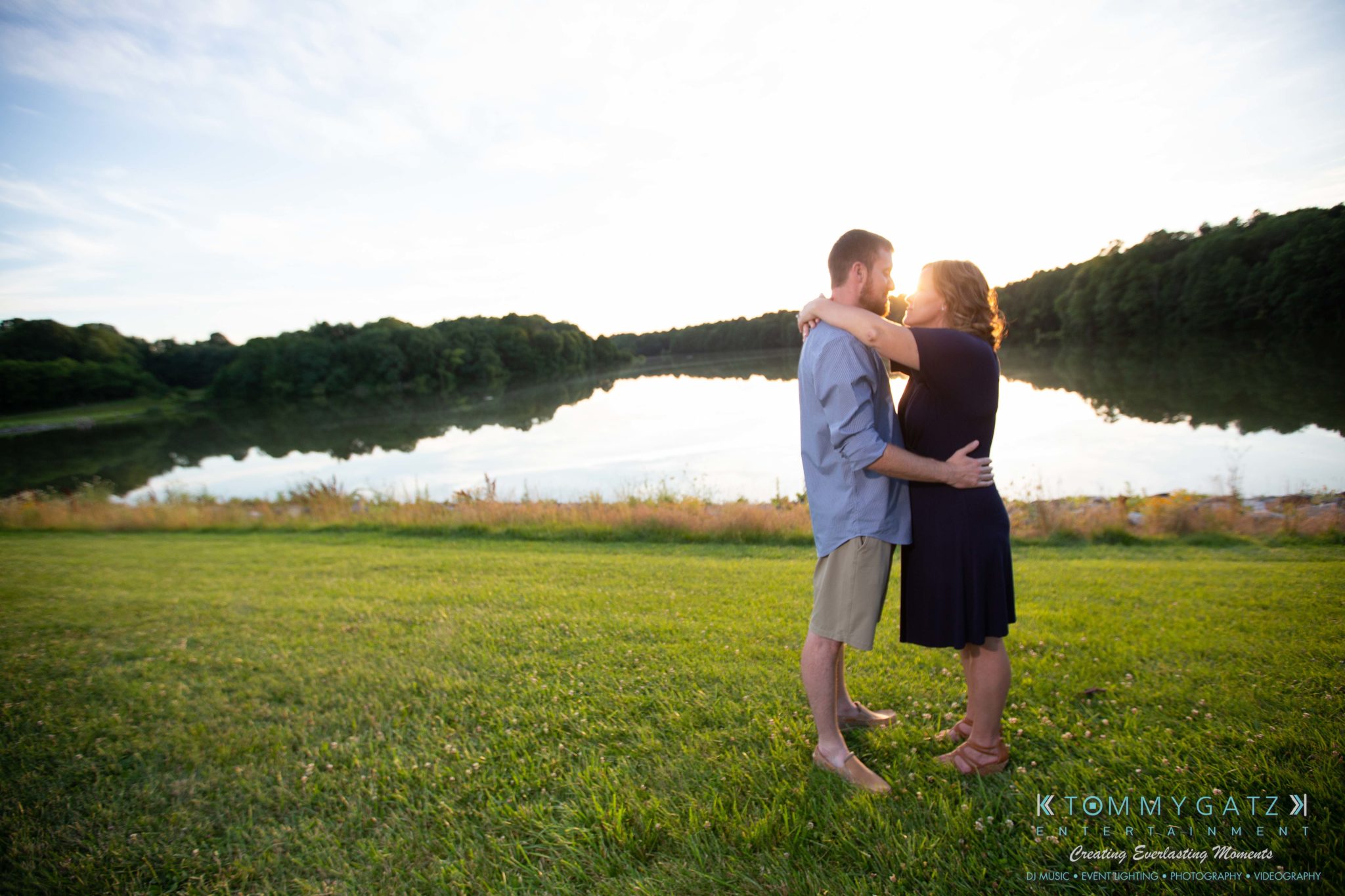 A couple embraces by a lake during sunset, standing on lush grass with trees lining the horizon and sunlight reflecting off the water.