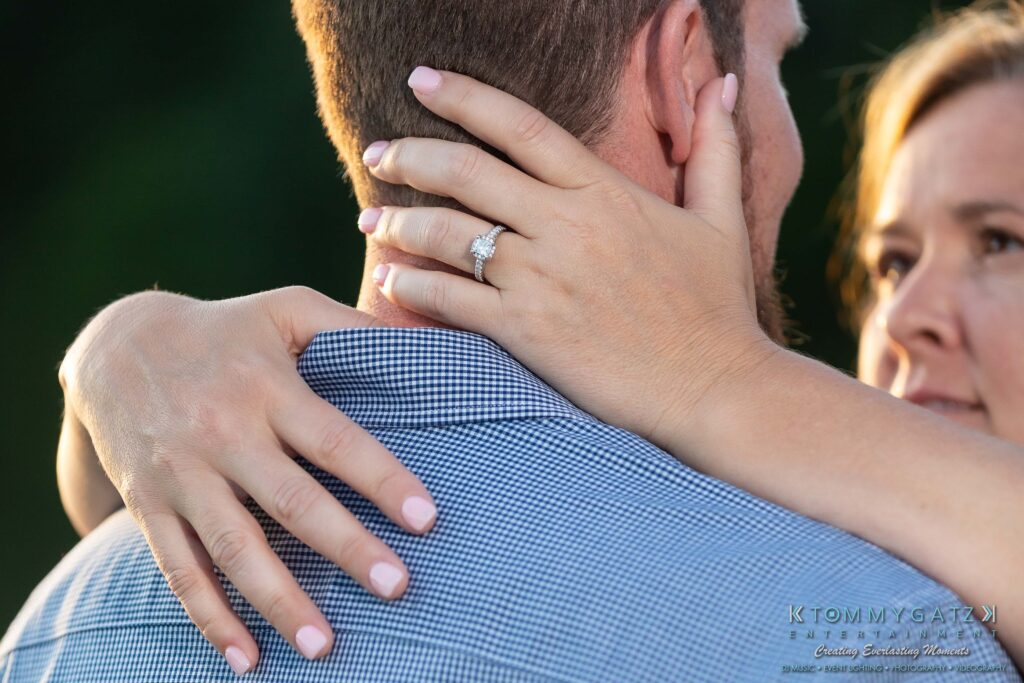 Heather-Scott-Engagement-Shoot-Low-Res-WM-43 - TOMMY GATZ ENTERTAINMENT Woman with engagement ring embraces a man, hands around his neck, both in casual attire, outdoors.