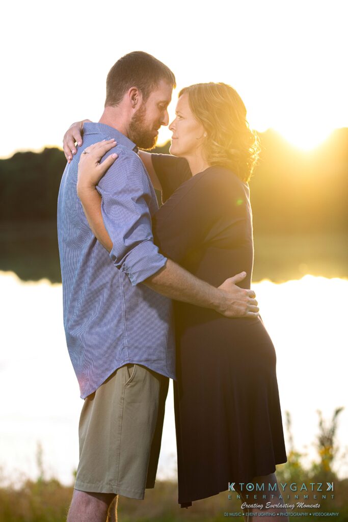 Heather-Scott-Engagement-Shoot-Low-Res-WM-44 - TOMMY GATZ ENTERTAINMENT A couple embraces by a lake at sunset, with the sun glowing behind them.