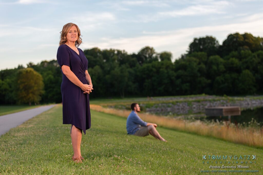 Heather-Scott-Engagement-Shoot-Low-Res-WM-46 - TOMMY GATZ ENTERTAINMENT A woman in a blue dress stands on a grassy area near a body of water, with a man sitting in the background. Trees and a cloudy sky are visible.