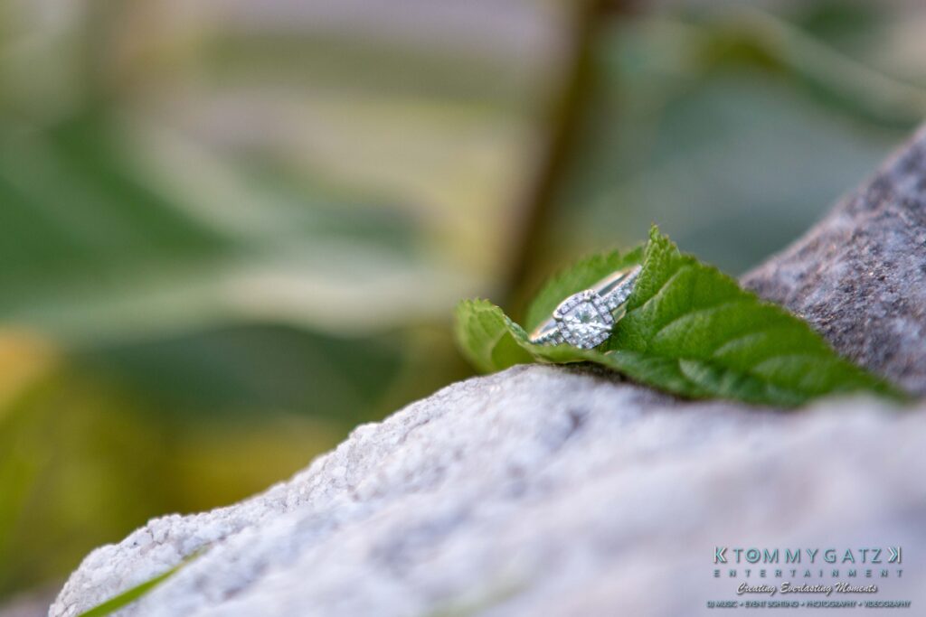 Heather-Scott-Engagement-Shoot-Low-Res-WM-48 - TOMMY GATZ ENTERTAINMENT Diamond engagement ring on a green leaf over a rock, with a blurred natural background.