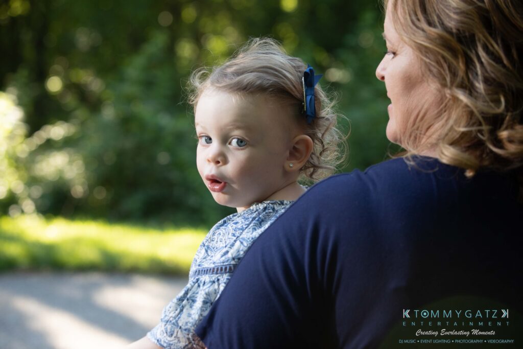 Heather-Scott-Engagement-Shoot-Low-Res-WM-7 - TOMMY GATZ ENTERTAINMENT A toddler with a blue bow in her hair being held by an adult outdoors in a park setting.