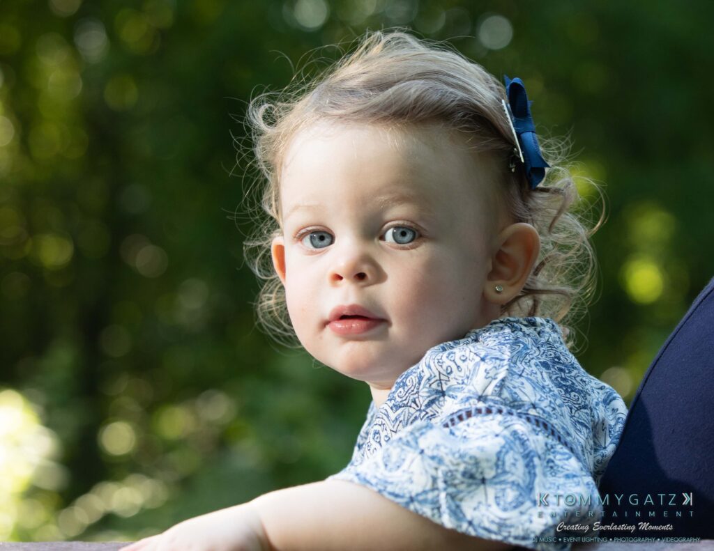 Heather-Scott-Engagement-Shoot-Low-Res-WM-8 - TOMMY GATZ ENTERTAINMENT Young child with curly hair and a blue bow, wearing a patterned shirt, looking towards the camera with trees in the background.