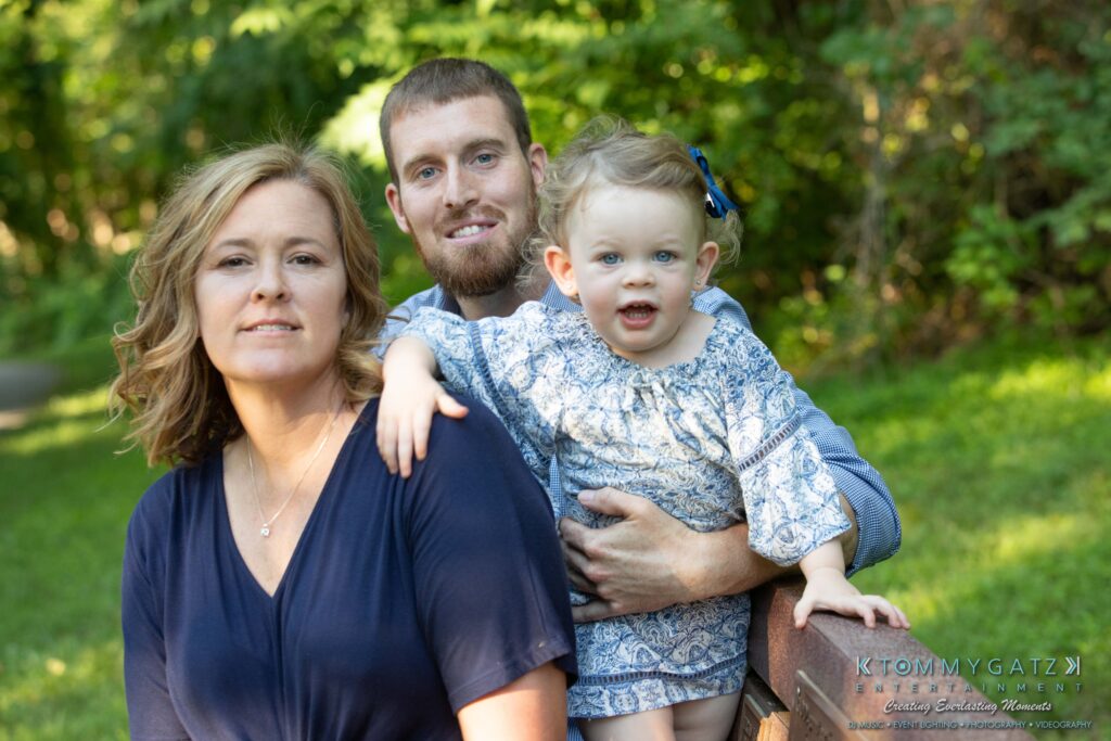 Heather-Scott-Engagement-Shoot-Low-Res-WM-9 - TOMMY GATZ ENTERTAINMENT A woman, man, and child pose outdoors on a bench. The child, wearing a blue patterned dress, is in front, with the adults alongside. Greenery is visible in the background.