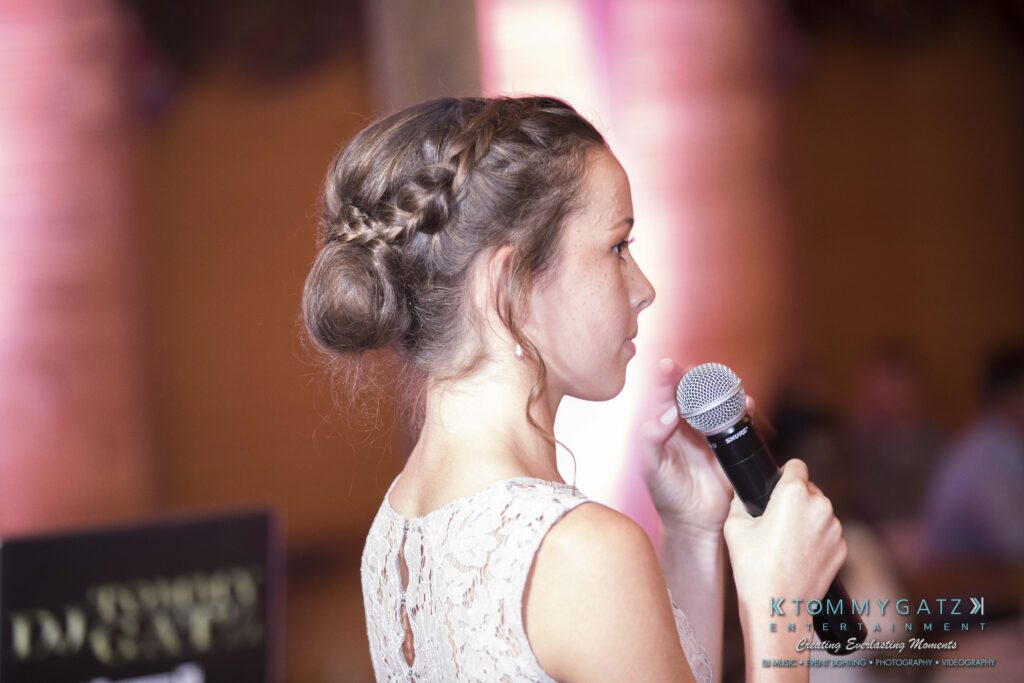 Jennifer-Rudi-Highlights-Low-res-100 - TOMMY GATZ ENTERTAINMENT A woman in a lace dress with braided hair faces sideways while holding a microphone.