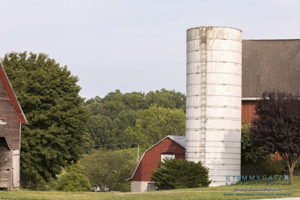 Jennifer-Rudi-Highlights-Low-res-5 - TOMMY GATZ ENTERTAINMENT Farm scene with a large white silo, red barn, and trees. The background has a line of lush green trees under a clear sky.