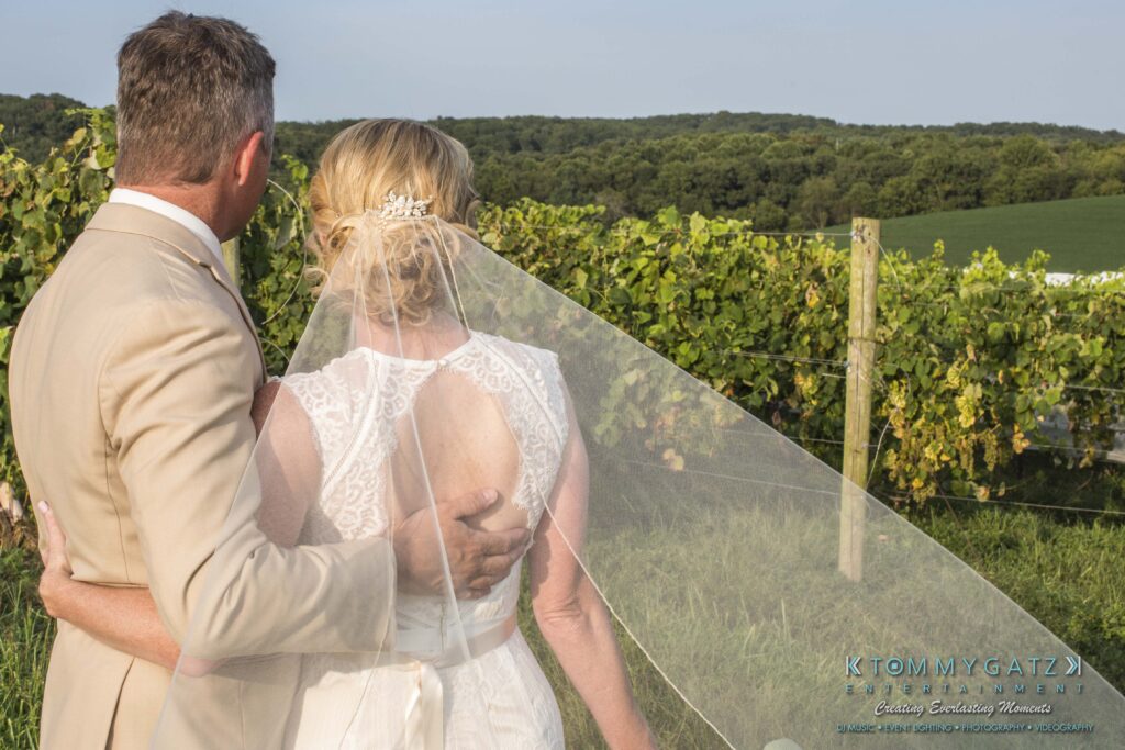 Jennifer-Rudi-Highlights-Low-res-51 - TOMMY GATZ ENTERTAINMENT A couple in wedding attire stands with their backs to the camera, looking at a vineyard. The man is in a beige suit and the woman is in a white dress with a veil.