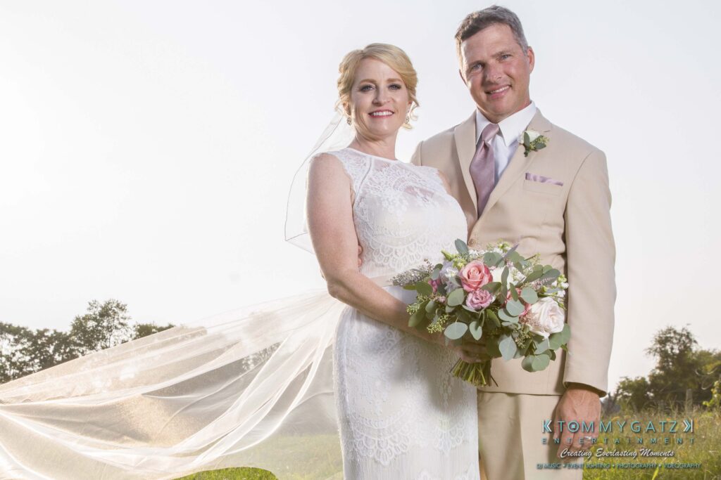 Jennifer-Rudi-Highlights-Low-res-52 - TOMMY GATZ ENTERTAINMENT A couple in wedding attire stands outdoors smiling, with the bride holding a bouquet of pink and white flowers.