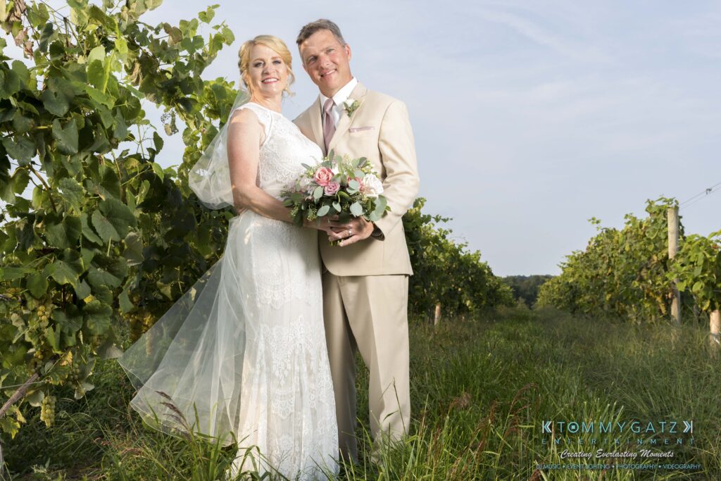 Jennifer-Rudi-Highlights-Low-res-54 - TOMMY GATZ ENTERTAINMENT A couple in wedding attire poses in a vineyard, with the woman holding a bouquet of pink and white flowers.
