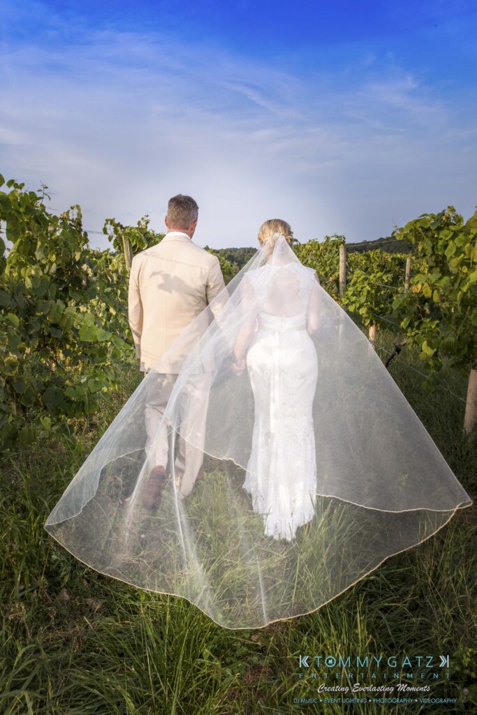Jennifer-Rudi-Highlights-Low-res-58 - TOMMY GATZ ENTERTAINMENT A bride and groom walk hand in hand through a vineyard, both dressed in formal attire, with the bride's long veil trailing behind them.