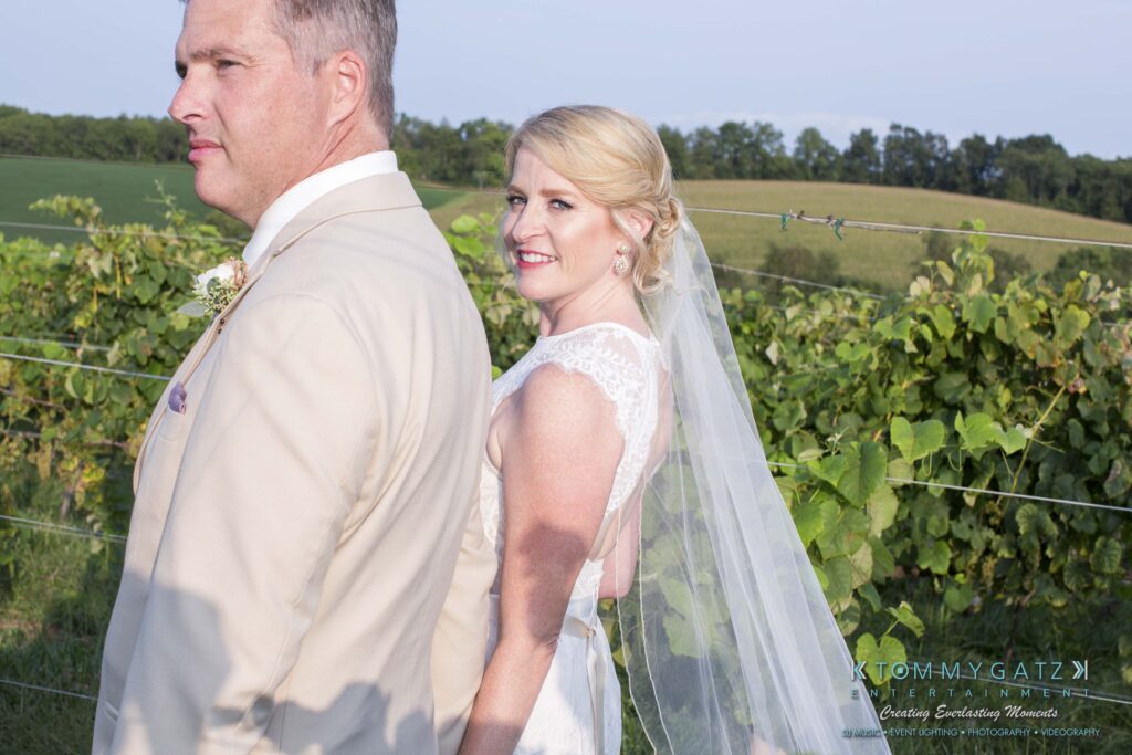 Jennifer-Rudi-Highlights-Low-res-60 - TOMMY GATZ ENTERTAINMENT A bride and groom stand back-to-back in a vineyard, with the bride smiling and the groom facing away. Both are in formal wedding attire.