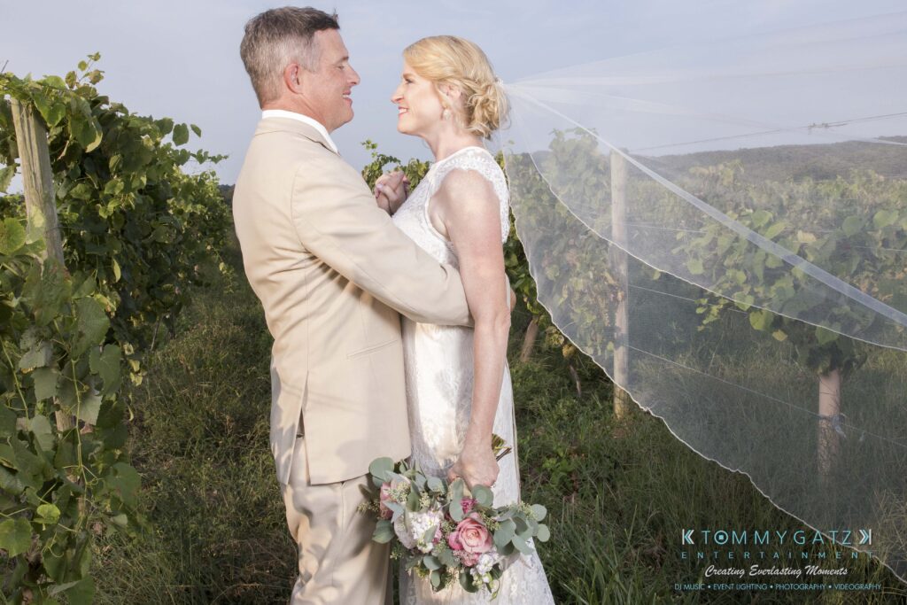 Jennifer-Rudi-Highlights-Low-res-62 - TOMMY GATZ ENTERTAINMENT A couple in wedding attire stands smiling in a vineyard, with the bride holding a bouquet and wearing a flowing veil.