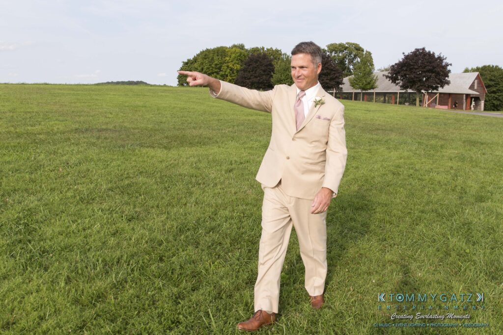 Jennifer-Rudi-Highlights-Low-res-7 - TOMMY GATZ ENTERTAINMENT A man in a beige suit with a pink tie gestures and smiles while standing on a grassy field with trees and a building in the background.