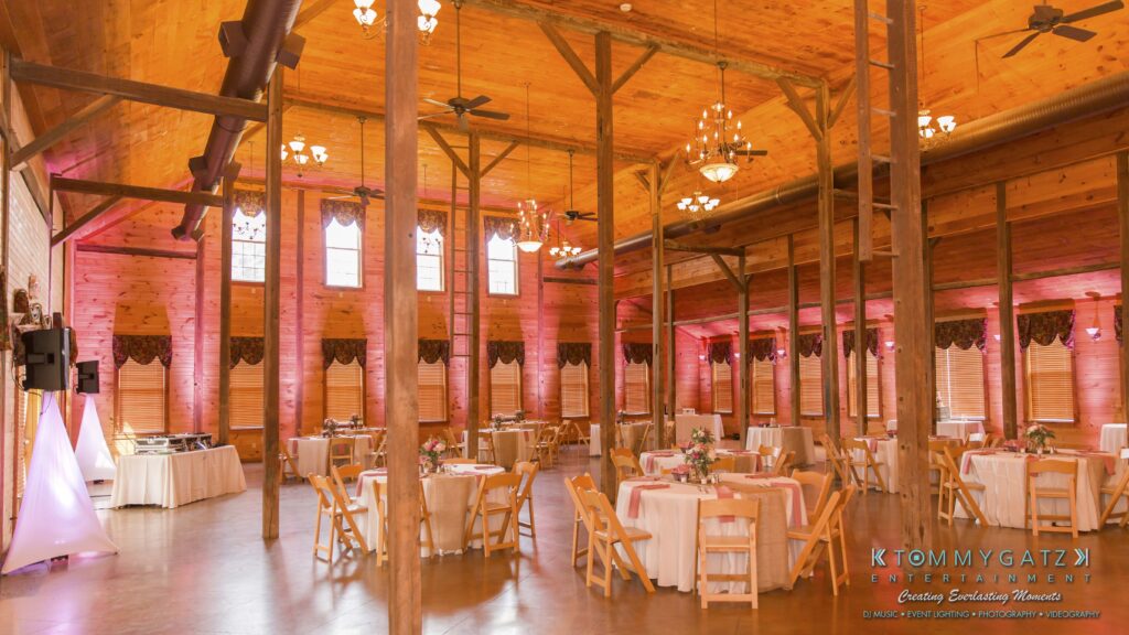 Jennifer-Rudi-Highlights-Low-res-79 - TOMMY GATZ ENTERTAINMENT A spacious, rustic-style banquet hall with wooden beams and chandeliers. Tables are set with white tablecloths and pink accents. Large windows let in natural light.