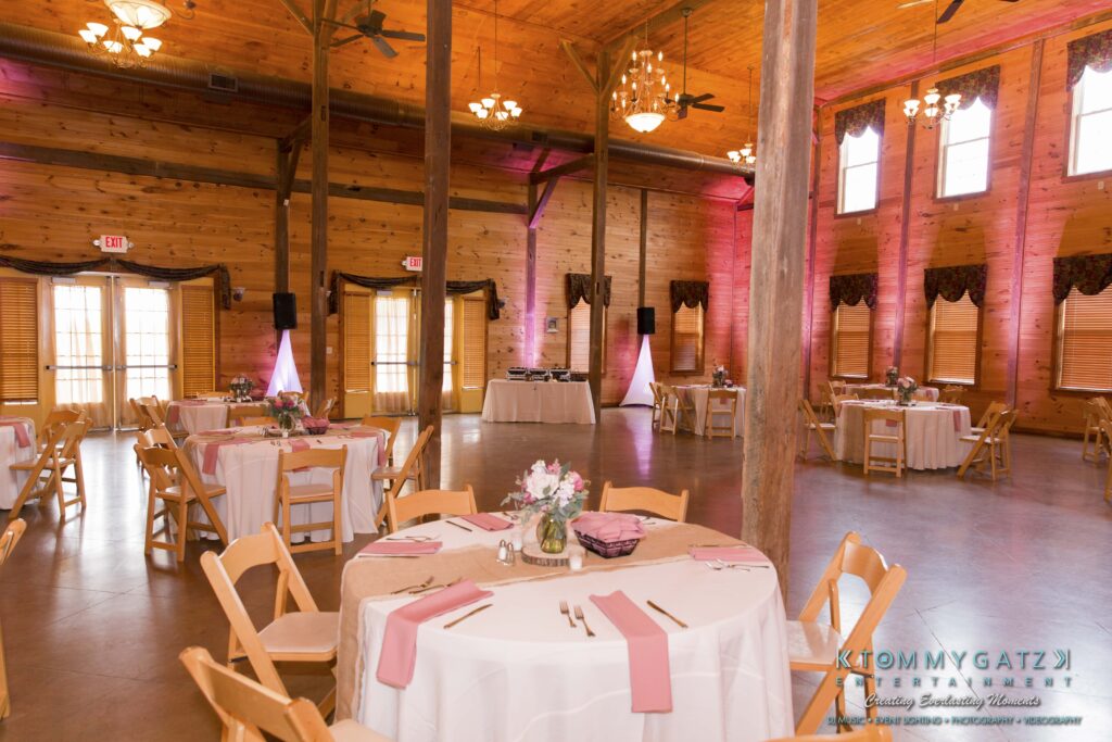 Jennifer-Rudi-Highlights-Low-res-80 - TOMMY GATZ ENTERTAINMENT Wooden event hall with round tables covered in white tablecloths and set with pink napkins. Floral centerpieces adorn the tables, and soft lighting creates a warm atmosphere.