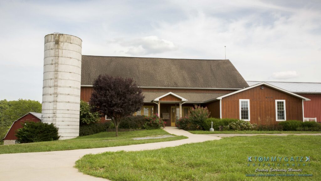 Jennifer-Rudi-Highlights-Low-res-83 - TOMMY GATZ ENTERTAINMENT A large barn with a grey silo and wooden exterior surrounded by greenery, under a partly cloudy sky.