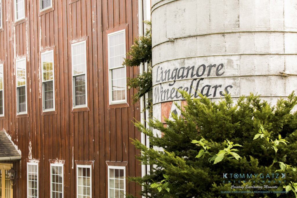 Jennifer-Rudi-Highlights-Low-res-84 - TOMMY GATZ ENTERTAINMENT Wooden building beside a large white silo with "Lingonore Winecellars" written on it, partially obscured by greenery.