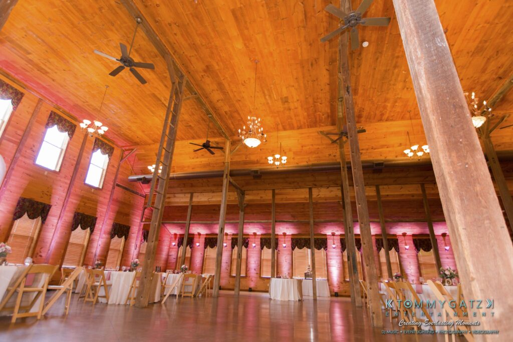 Jennifer-Rudi-Highlights-Low-res-86 - TOMMY GATZ ENTERTAINMENT Large rustic wooden hall with high ceiling, visible beams, ceiling fans, and long windows. Decor includes tables with white cloths and pink uplighting.