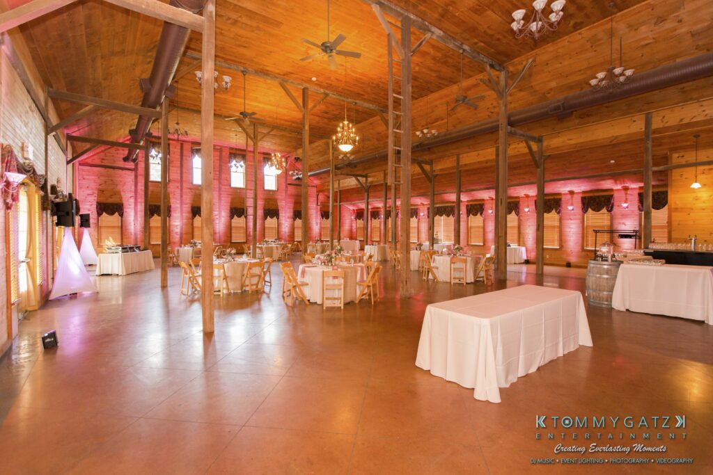 Jennifer-Rudi-Highlights-Low-res-87 - TOMMY GATZ ENTERTAINMENT Spacious wooden event hall with round tables set for a gathering. Chandeliers hang from the ceiling, and the room features large windows and subdued lighting.