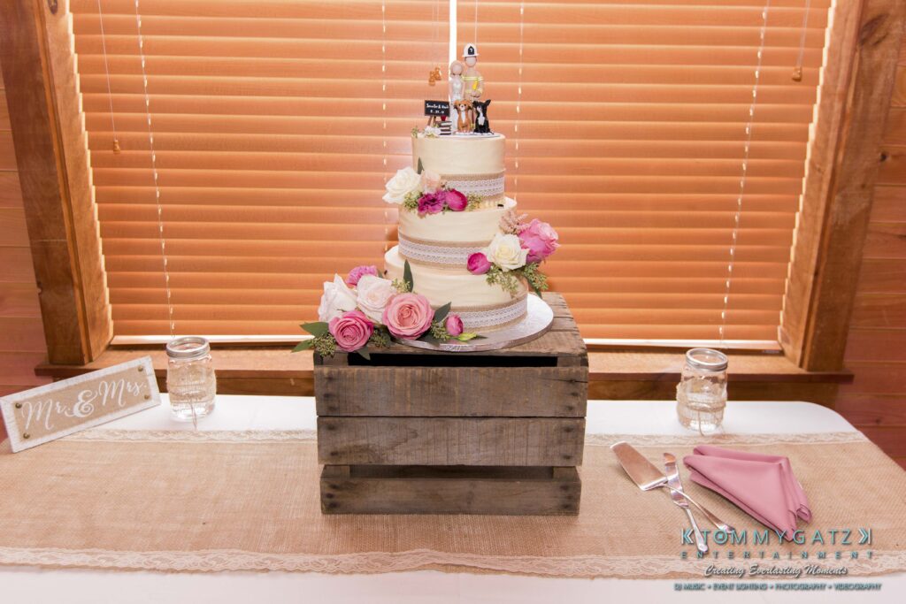 Jennifer-Rudi-Highlights-Low-res-88 - TOMMY GATZ ENTERTAINMENT A three-tiered wedding cake with floral decorations sits on a wooden crate. A "Mr. & Mrs." sign is on the table, along with pink napkins and utensils.