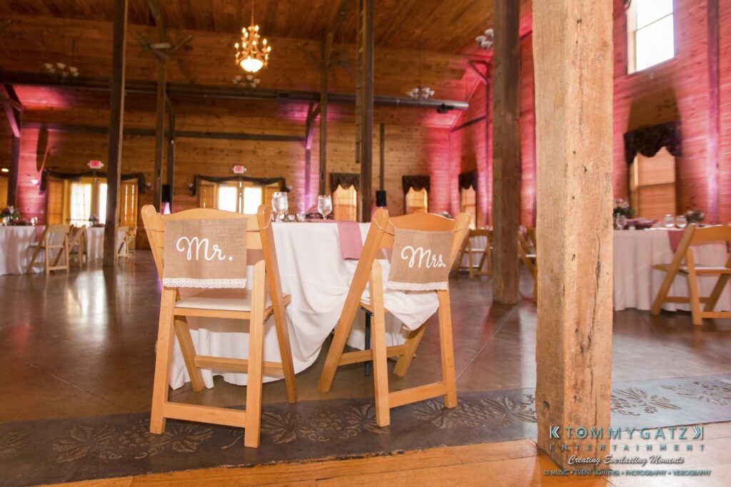 Jennifer-Rudi-Highlights-Low-res-89 - TOMMY GATZ ENTERTAINMENT Wooden wedding reception hall with round tables, white tablecloths, and chairs labeled "Mr." and "Mrs." Pink lighting and rustic wooden beams create a warm atmosphere.