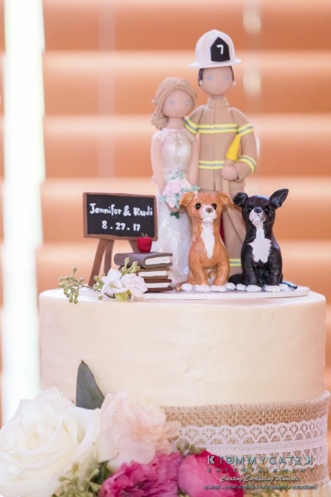 Jennifer-Rudi-Highlights-Low-res-94 - TOMMY GATZ ENTERTAINMENT Wedding cake with a firefighter and bride topper, a small chalkboard with "Jennifer & Rudi 8.27.17," two dog figurines, and flowers at the base.