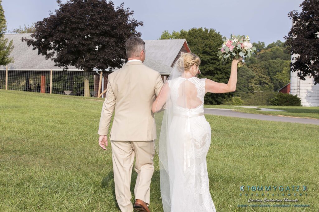 JenniferRudiWeddingCeremonyPhotos-174-1 - TOMMY GATZ ENTERTAINMENT A couple in formal attire walks on grass, with the woman holding a bouquet. They are headed toward a barn and trees in the background.