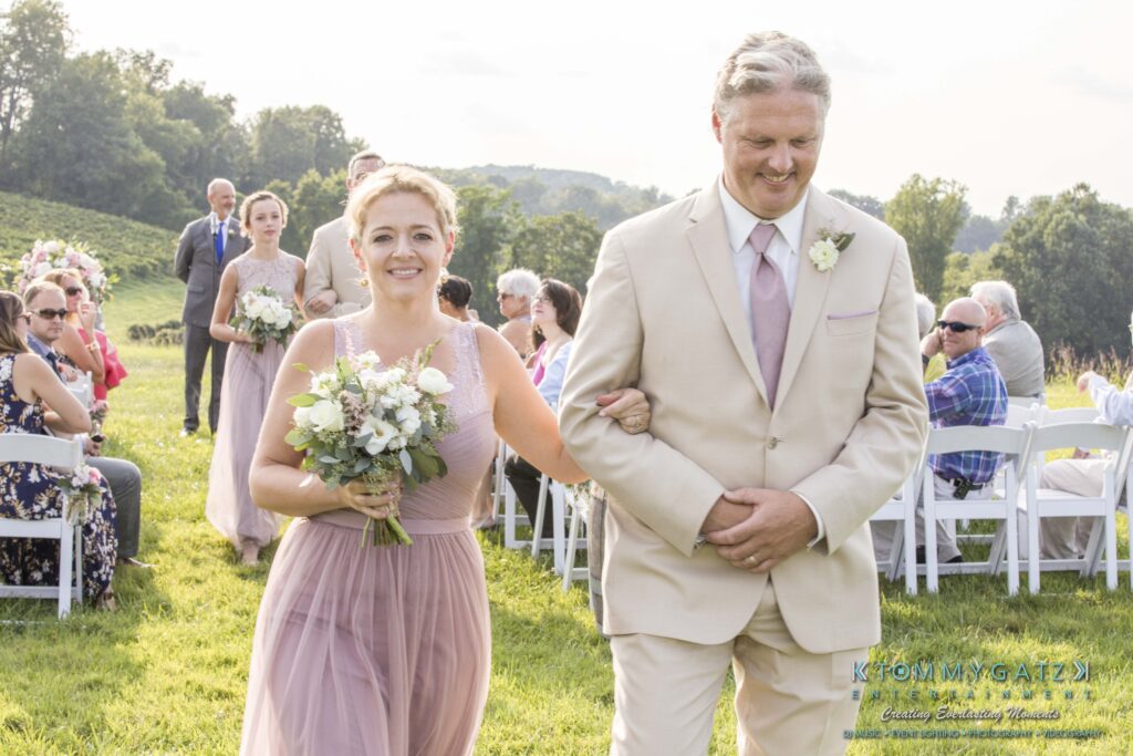 JenniferRudiWeddingCeremonyPhotos-175 - TOMMY GATZ ENTERTAINMENT A woman and man in formal attire walk down an outdoor aisle. She holds a bouquet; guests are seated on either side.