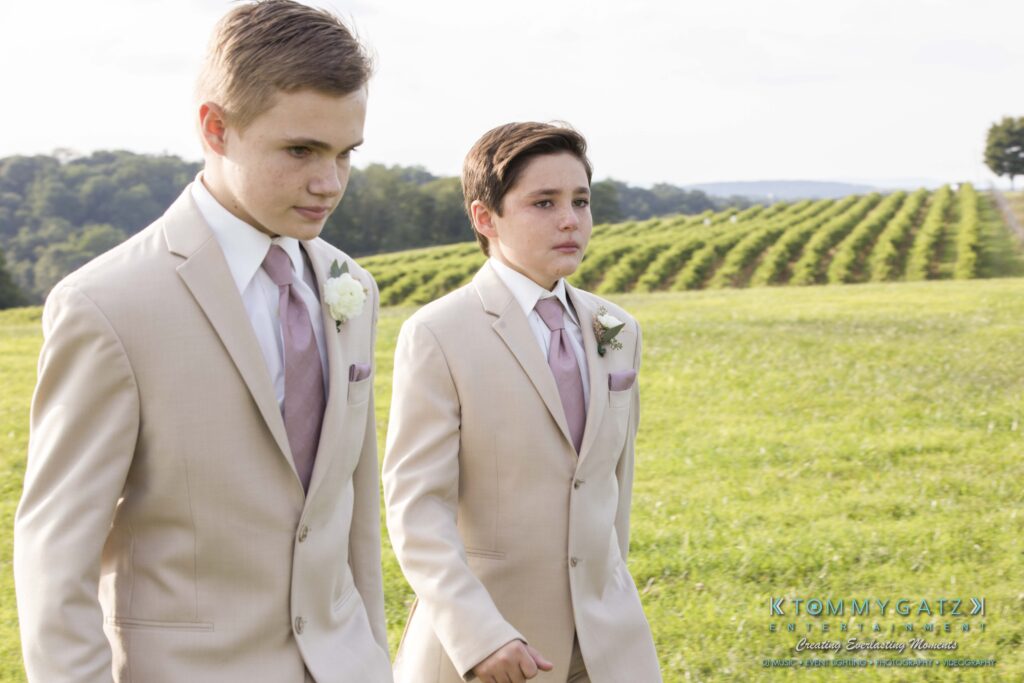 JenniferRudiWeddingCeremonyPhotos-177 - TOMMY GATZ ENTERTAINMENT Two young boys in cream suits and lavender ties walk through a grassy field with vineyards in the background.