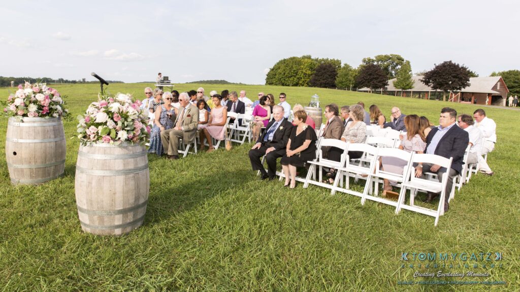 JenniferRudiWeddingCeremonyPhotos-70 - TOMMY GATZ ENTERTAINMENT A group of people seated on white chairs at an outdoor event, surrounded by green grass and floral barrel decorations.