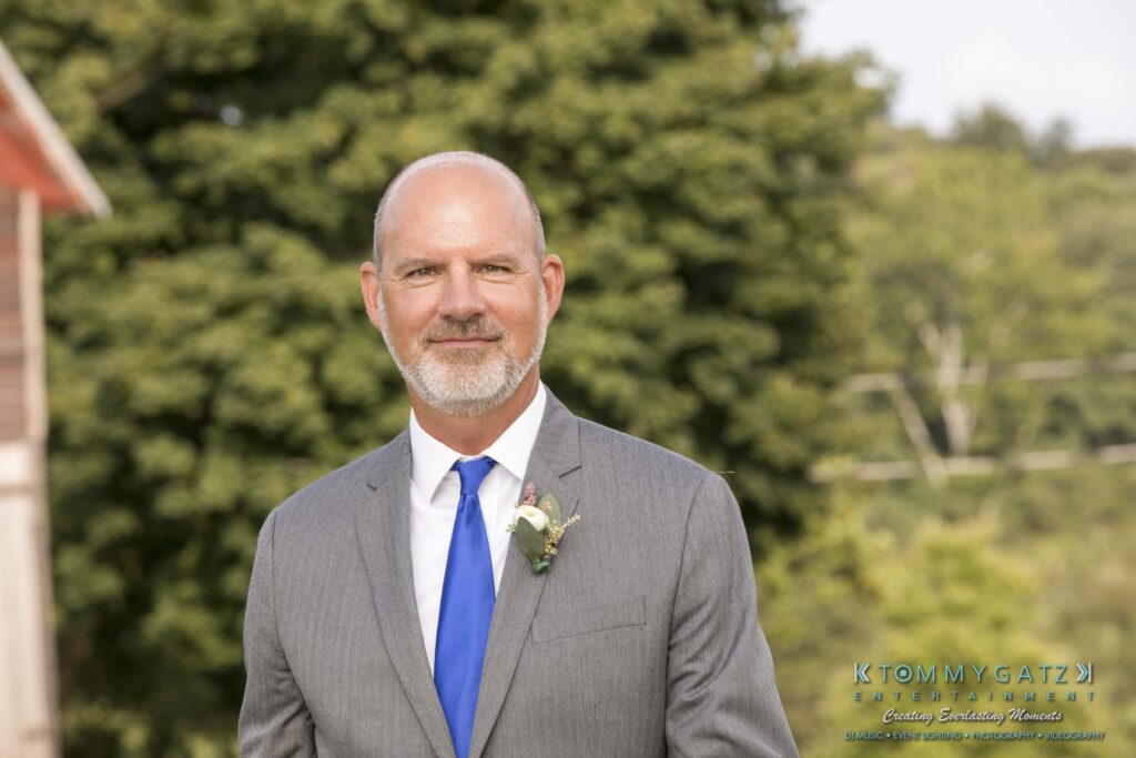 JenniferRudiWeddingCeremonyPhotos-72 - TOMMY GATZ ENTERTAINMENT Man in a gray suit and blue tie stands outdoors with trees in the background.