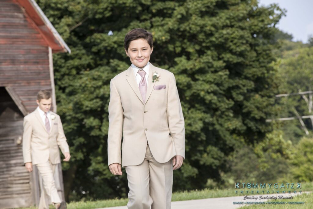 JenniferRudiWeddingCeremonyPhotos-73 - TOMMY GATZ ENTERTAINMENT A young boy in a beige suit with a pink tie and boutonnière walks outdoors. Another boy in a similar outfit follows. Trees and a barn are in the background.